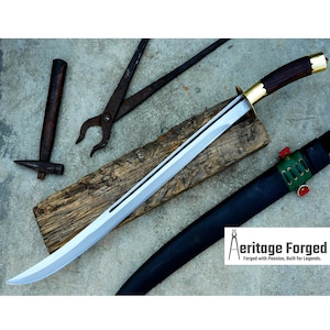 May include: A long, silver-colored sword with a dark wood handle and gold accents. The sword is lying on a wooden surface with a black leather sheath and a hammer and pliers in the background. The text "Heritage Forged" is visible in the lower right corner of the image.