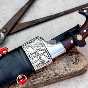May include: A black leather-wrapped knife with a silver handle featuring an engraved design. The knife has a wooden handle and a sharp blade. The knife is resting on a piece of wood.