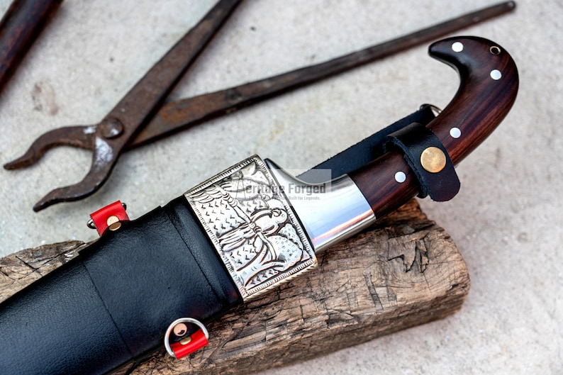May include: A black leather-wrapped knife with a silver handle featuring an engraved design. The knife has a wooden handle and a sharp blade. The knife is resting on a piece of wood.