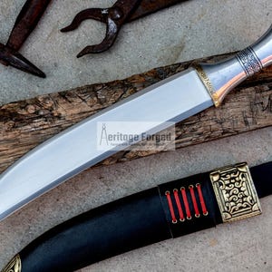 May include: A silver and gold dagger with a curved blade and a decorative handle. The dagger is resting on a piece of wood and has a black leather sheath with a gold buckle.