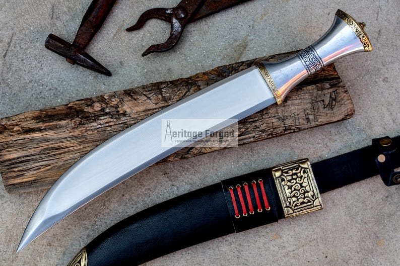 May include: A silver and gold dagger with a curved blade and a decorative handle. The dagger is resting on a piece of wood and has a black leather sheath with a gold buckle.