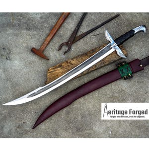 Könnte beinhalten: Ein handgeschmiedetes Schwert mit silberner Klinge und schwarzem Griff, das auf einem Holzbrett liegt. Das Schwert wird von einer weinroten Lederscheide mit grünen Akzenten begleitet. Schmiedewerkzeuge sind im Hintergrund zu sehen. Der Text "Aeritage Forged" befindet sich unten rechts.