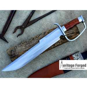 May include: A large, polished steel blade with a wooden handle and a guard. The blade rests on a wooden plank, with a brown leather sheath and the text "Aeritage Forged" visible. Other tools are in the background.