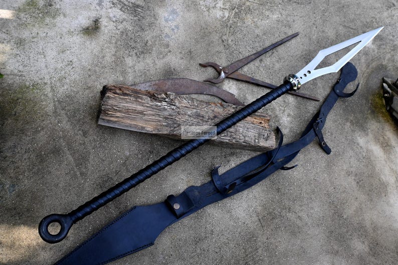 Puede incluir: Una lanza negra y plateada con una funda de cuero y un mango de madera. La lanza tiene una hoja en forma de diamante y un anillo de metal decorativo en el extremo del mango.