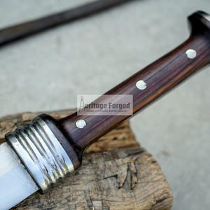 May include: A close-up of a knife with a dark wood handle and a silver blade. The handle has a decorative silver band and two silver dots. The text "Heritage Forged" is visible on the handle.