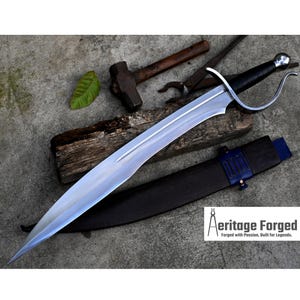 May include: A silver sword with a black leather handle and a blue scabbard. The sword has a curved blade and a decorative pommel. The sword is lying on a wooden surface with a hammer and other tools.