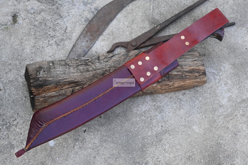 May include: A handcrafted leather sheath in deep purple, with a burgundy belt and brass rivets. The sheath is designed to hold a blade, resting on a weathered wooden block. The background includes antique tools.