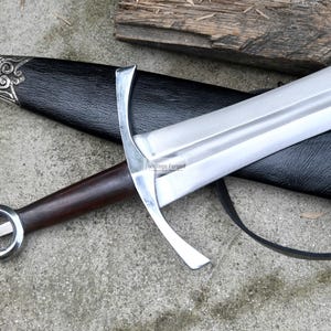 Op de afbeelding: Een zilveren en bruin zwaard met een zwarte leren schede. Het zwaard heeft een decoratief handvat en een lang, scherp blad.