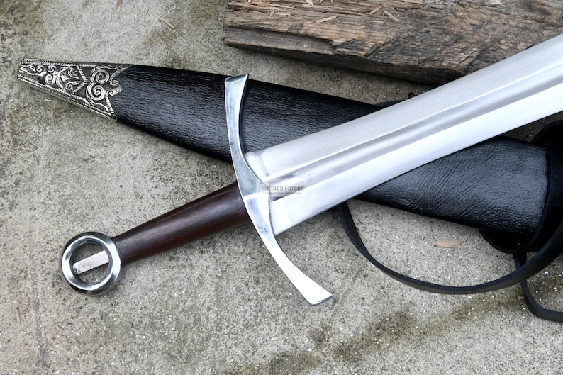 Op de afbeelding: Een zilveren en bruin zwaard met een zwarte leren schede. Het zwaard heeft een decoratief handvat en een lang, scherp blad.