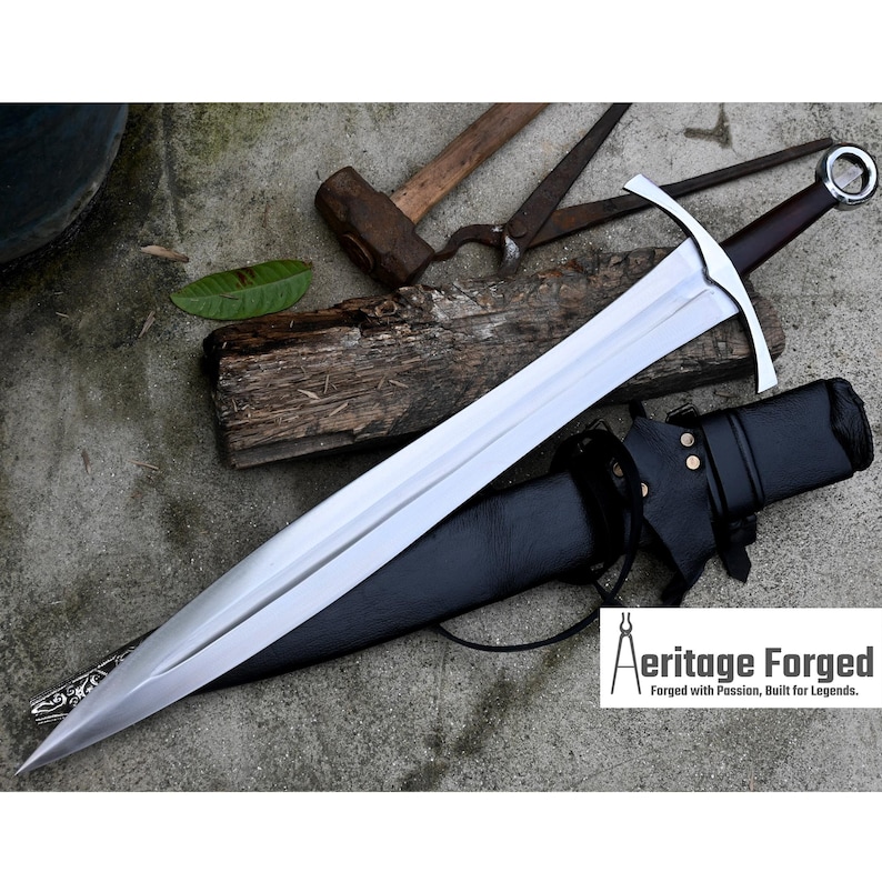 Op de afbeelding: Een zilveren zwaard met een zwarte leren schede en een houten handvat. Het zwaard ligt op een grijs oppervlak met een hamer, tang en een stuk hout op de achtergrond. De tekst "Heritage Forged" is zichtbaar in de rechterbenedenhoek van de afbeelding.  Forged with Passion, Built for Legends.