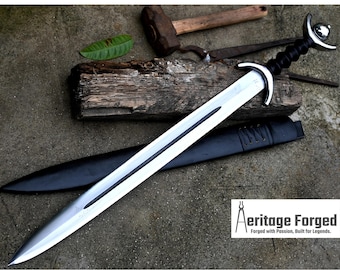 May include: A silver sword with a black handle and a leather scabbard. The sword has a crescent-shaped guard and a pointed tip. The sword is resting on a wooden surface. The text "Heritage Forged" is visible in the lower right corner of the image. "Forged with Passion, Built for Legends."