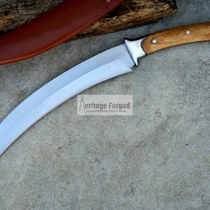 May include: A curved, silver-colored knife with a wooden handle. The handle has a brown wood grain and two silver rivets. The knife is on a gray concrete surface. The text "Heritage Forged" is visible in the image.