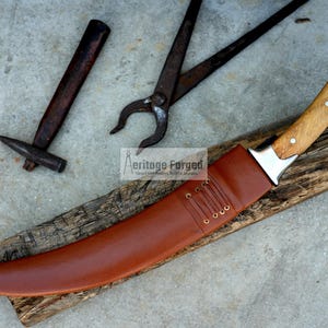 May include: A curved, brown leather-sheathed knife with a wooden handle and a silver blade. The knife is resting on a piece of wood with a hammer and a pair of pliers in the background. The text "Heritage Forged" is visible in the image.