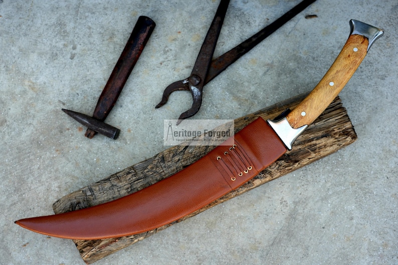 May include: A curved, brown leather-sheathed knife with a wooden handle and a silver blade. The knife is resting on a piece of wood with a hammer and a pair of pliers in the background. The text "Heritage Forged" is visible in the image.