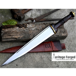 May include: A long, silver-colored dagger with a dark brown wooden handle and gold accents. The dagger is resting on a brown leather sheath with a decorative pattern. The text "Heritage Forged" is visible in the lower right corner of the image.  Forged with Passion, Built for Legends.