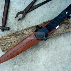 May include: A brown leather sheath with a black leather strap and a silver metal buckle. The sheath is holding a long, silver blade with a black handle. The blade is sharp and pointed. The sheath is resting on a piece of wood. The text "Heritage Forged" is visible on the sheath.