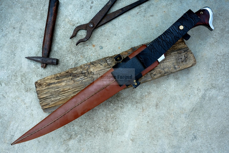 May include: A brown leather sheath with a black leather strap and a silver metal buckle. The sheath is holding a long, silver blade with a black handle. The blade is sharp and pointed. The sheath is resting on a piece of wood. The text "Heritage Forged" is visible on the sheath.