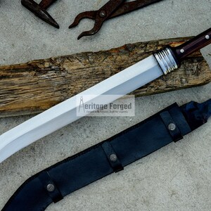 May include: A silver and brown curved sword with a wooden handle and a black leather sheath. The sword is on a wooden surface and the sheath is on a gray surface. The text "Heritage Forged" is visible on the blade.