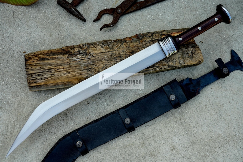 May include: A silver and brown curved sword with a wooden handle and a black leather sheath. The sword is on a wooden surface and the sheath is on a gray surface. The text "Heritage Forged" is visible on the blade.