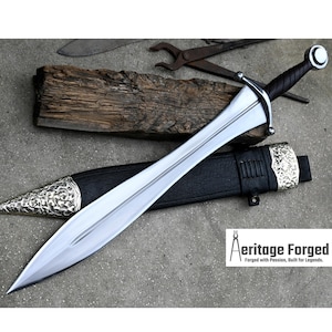 May include: A silver and black sword with a leather scabbard. The sword has a decorative handle and a pointed blade. The scabbard has a silver metal tip and a decorative design. The sword is on a wooden surface. The text "Heritage Forged" is visible in the lower right corner of the image. "Forged with Passion, Built for Legends."