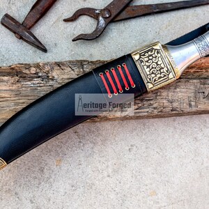 May include: A black leather-wrapped knife with a gold-colored handle and intricate designs. The handle has red stitching and a silver metal pommel. The knife is resting on a piece of weathered wood.
