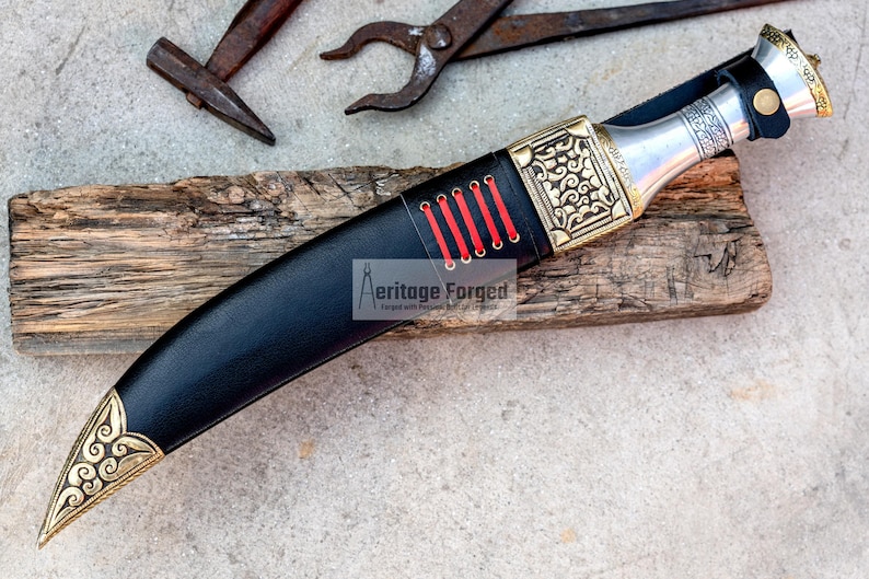 May include: A black leather-wrapped knife with a gold-colored handle and intricate designs. The handle has red stitching and a silver metal pommel. The knife is resting on a piece of weathered wood.