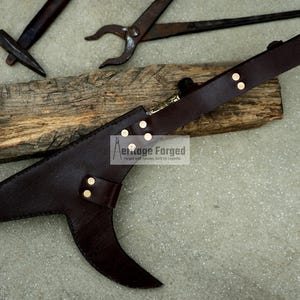 May include: A brown leather scabbard with a metal ring at the top. The scabbard has a curved blade shape and is decorated with gold rivets. The text "Heritage Forged" is visible on the scabbard.