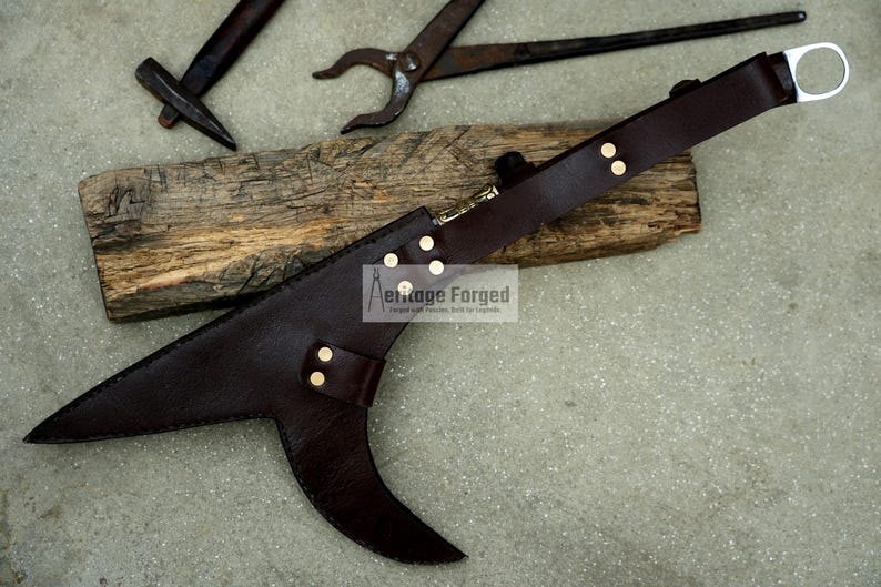 May include: A brown leather scabbard with a metal ring at the top. The scabbard has a curved blade shape and is decorated with gold rivets. The text "Heritage Forged" is visible on the scabbard.