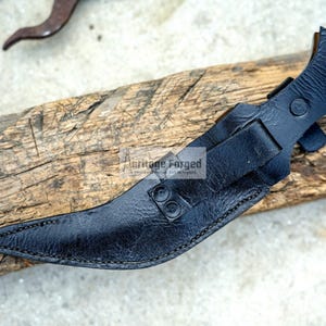 May include: Black leather knife sheath with a curved blade and a metal tip. The sheath has a decorative stitching detail and a metal button.