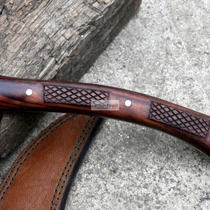 May include: Close-up of a handcrafted knife with a dark wooden handle. The handle features a carved diamond pattern and is secured with silver rivets. A leather sheath is partially visible.