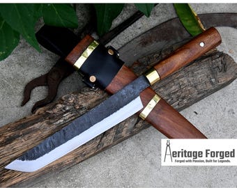 May include: A black leather sheath with a brass accent and a wooden handle is attached to a long, sharp, black blade. The blade is resting on a wooden surface. The text "Heritage Forged" is visible in the lower right corner of the image. "Forged with Passion, Built for Legends." is also visible.