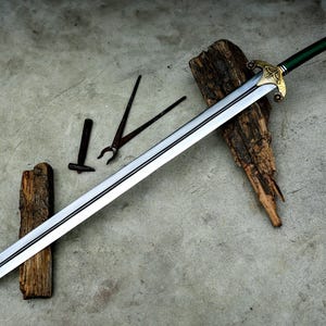 May include: A long, silver sword with a green handle and gold accents, resting on a weathered wooden surface. Blacksmith tools, including tongs and a hammer, are also visible. The sword's blade is approximately 36 inches long.