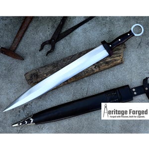 Könnte beinhalten: Ein langes Schwert mit polierter Silberklinge und dunklem Holzgriff mit kreisförmigem Handschutz. Das Schwert liegt auf einem Holzstück, darunter eine schwarze Lederscheide. Der Text "Aeritage Forged" ist sichtbar.