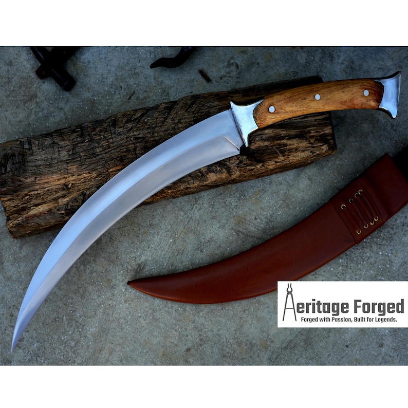 May include: A curved, silver-colored knife with a wooden handle. The knife is resting on a piece of wood and has a brown leather sheath.