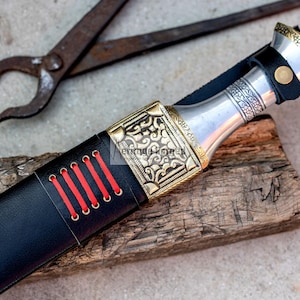 May include: A black leather knife sheath with red stitching and a silver and gold metal handle with intricate designs.