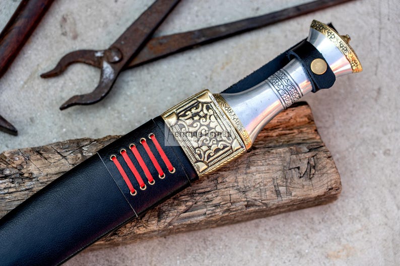 May include: A black leather knife sheath with red stitching and a silver and gold metal handle with intricate designs.