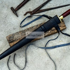 May include: A black dagger with a wooden handle and gold accents, resting in a black sheath. The dagger is on a wooden plank with a blue strap. Tools are in the background.