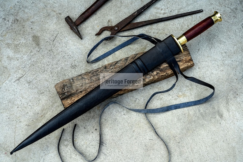 May include: A black dagger with a wooden handle and gold accents, resting in a black sheath. The dagger is on a wooden plank with a blue strap. Tools are in the background.