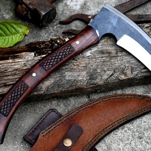 May include: A curved blade knife with a dark wooden handle featuring a diamond pattern. The blade is silver and white, and the knife rests on a weathered wooden surface. A brown leather sheath is also present.