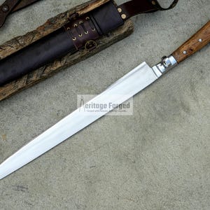 May include: A long, silver blade with a wooden handle. The blade is sharp and pointed, and the handle is made of a dark wood. The blade is lying on a gray surface. The text "Heritage Forged" is visible on the blade.