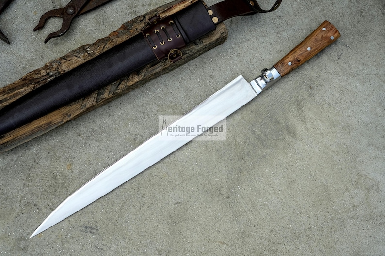 May include: A long, silver blade with a wooden handle. The blade is sharp and pointed, and the handle is made of a dark wood. The blade is lying on a gray surface. The text "Heritage Forged" is visible on the blade.