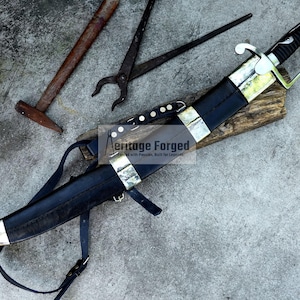 May include: A black and silver knife with a leather sheath and strap, resting on a wooden block. The knife has a curved blade and a decorative handle. The image also includes tools like a hammer and pliers. The text "Heritage Forged" is visible.