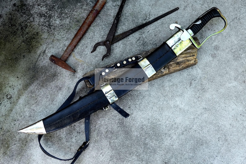 May include: A black and silver knife with a leather sheath and strap, resting on a wooden block. The knife has a curved blade and a decorative handle. The image also includes tools like a hammer and pliers. The text "Heritage Forged" is visible.