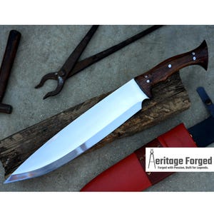 May include: A large, polished steel knife with a dark wood handle, resting on a wooden plank. The knife has a wide blade and is accompanied by a red leather sheath. Blacksmith tools are in the background.