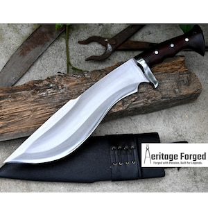 May include: A silver-colored, curved blade with a brown wooden handle. The blade is sharp and has a slight curve. The handle is made of wood and has a dark brown finish. The blade is resting on a piece of wood. The blade is in a black leather sheath with a brown leather strap. The sheath has a decorative stitching pattern. The text "Heritage Forged" is printed on the sheath. The text "Forged with Passion, Built for Legends." is printed below the logo.