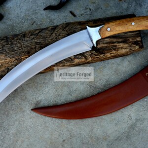 May include: A curved, silver-colored knife with a wooden handle and a brown leather sheath. The knife is resting on a piece of wood with the text "Heritage Forged" printed on it.