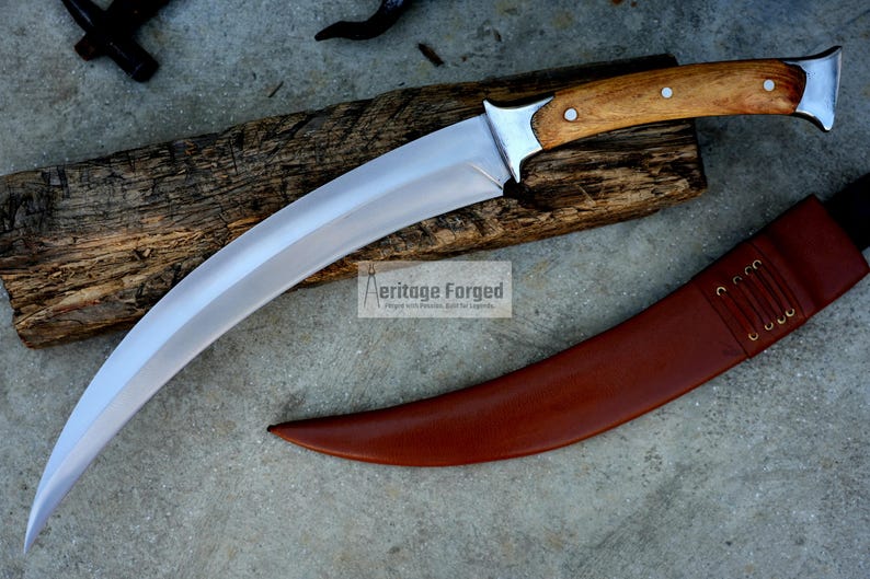 May include: A curved, silver-colored knife with a wooden handle and a brown leather sheath. The knife is resting on a piece of wood with the text "Heritage Forged" printed on it.