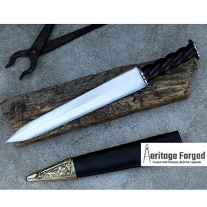 May include: A handmade, silver-bladed dagger with a dark, twisted wooden handle and a black leather sheath. The sheath has a decorative gold-colored metal end. The image also shows a pair of metal tongs and the text "Aeritage Forged".