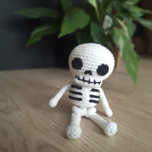Pode incluir: Boneca de esqueleto branca de crochê com detalhes pretos nos olhos, boca e caixa torácica. A boneca feita à mão está sentada em uma superfície de madeira. Os braços e pernas da boneca estão estendidos.