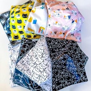 May include: A set of eight fabric bowl cozies featuring various cat designs. The cozies are made of cotton fabric and have a quilted design. The designs include black cats, white cats, and orange cats.
